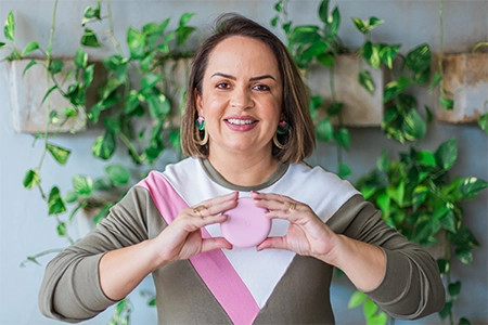 foto da Priscila Pagioro sorrindo, de pé, com uma blusa branca com rosa e marrom, segurando uma caixa rosa de invisalign
